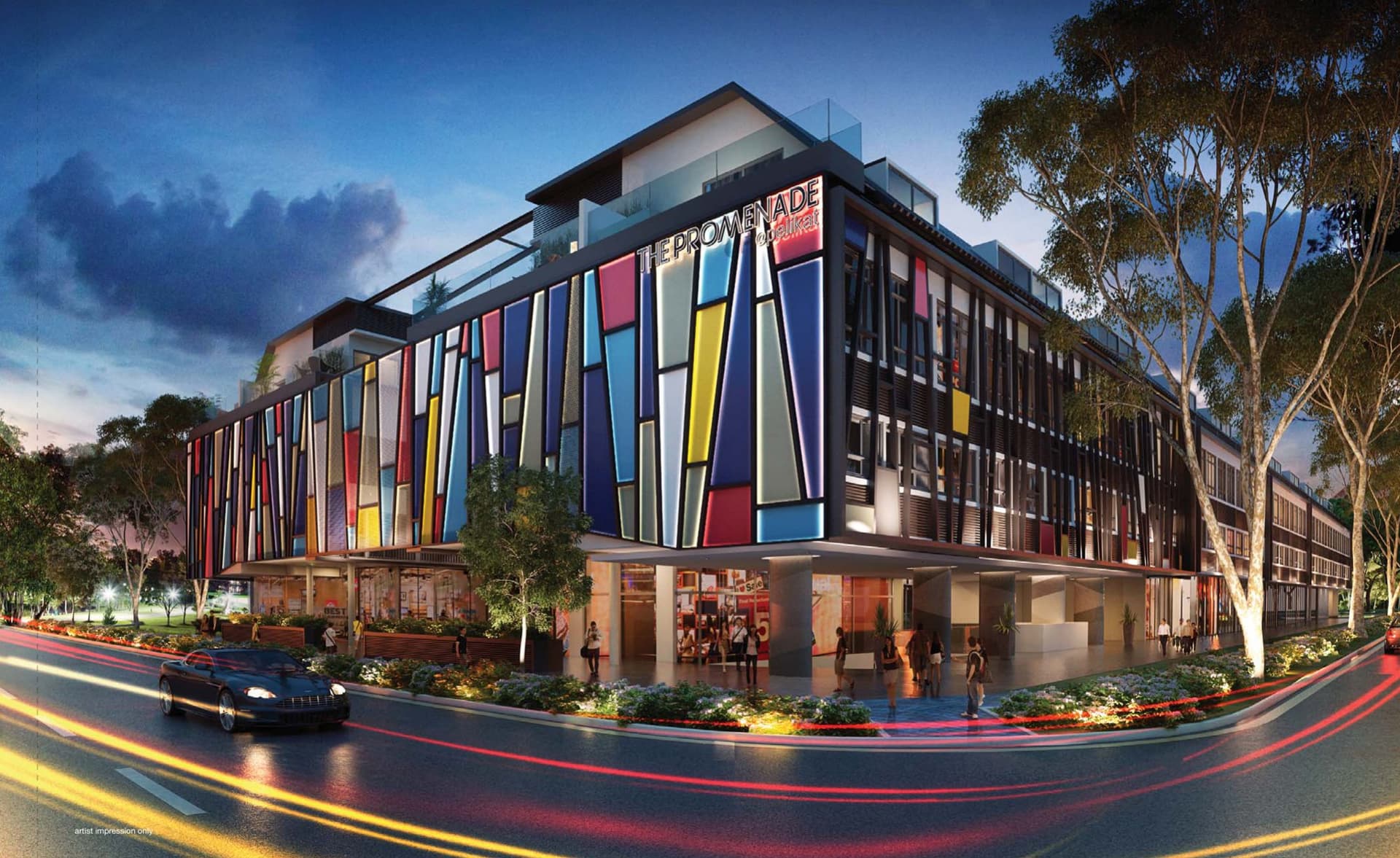 The Promenade @ Pelikat at dusk: a four-storey corner building with a coloured glass facade and shops on the ground floor along Jalan Pelikat